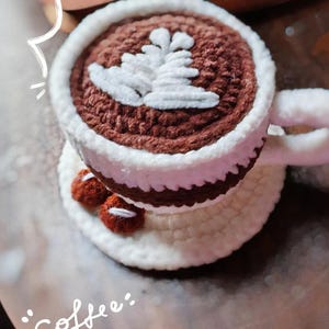 May include: Crocheted coffee cup with a white rim and handle, filled with brown yarn and topped with a white leaf design. The cup sits on a white saucer with two small, red crocheted beans. The words "coffee time" are written in white.