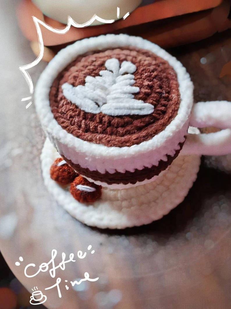 May include: Crocheted coffee cup with a white rim and handle, filled with brown yarn and topped with a white leaf design. The cup sits on a white saucer with two small, red crocheted beans. The words "coffee time" are written in white.