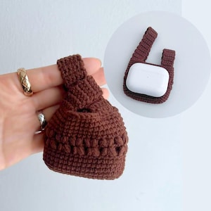Puede incluir: Una bolsa marrón tejida a mano con un nudo en la parte superior, diseñada para guardar objetos pequeños. La bolsa se muestra sostenida por una mano, con un anillo dorado visible. Una segunda bolsa similar contiene un dispositivo electrónico blanco.