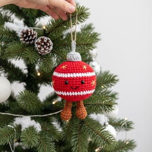 Peut inclure: Une boule de No&euml;l crochet&eacute;e &agrave; la main, rouge et blanche, avec un visage souriant et de petites jambes marron. L'ornement a un dessus gris et est d&eacute;cor&eacute; d'&eacute;clats d'&eacute;toiles jaunes. Il est suspendu &agrave; une ficelle en ficelle naturelle sur un sapin de No&euml;l.