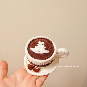 May include: A crocheted coffee cup and saucer. The cup is brown with a white leaf design and a white rim. The saucer has two small crocheted cookies. Text reads: "Come for a cup of coffee." and "Drink more coffee and talk less."
