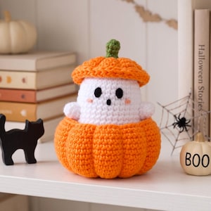 May include: A crochet amigurumi figure featuring a white ghost with black eyes and a small mouth, wearing an orange pumpkin hat and sitting inside a larger orange pumpkin base with a green stem on top of the hat.