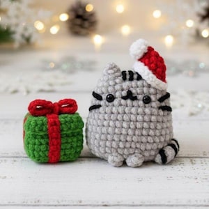 Op de afbeelding: Een gehaakte grijze kat met een rode en witte kerstmuts naast een groene geschenkdoos met een rode strik. De kat heeft zwarte snorharen en een gestreepte staart. De geschenkdoos heeft een rood lint.