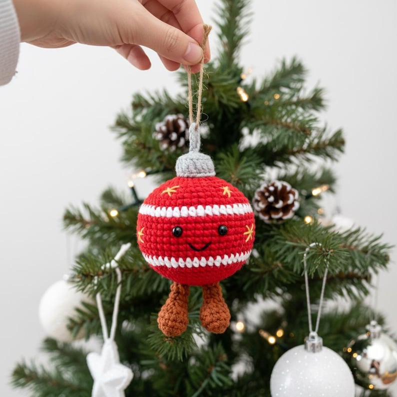 Peut inclure: Une d&eacute;coration de No&euml;l crochet&eacute;e rouge et blanche avec un visage souriant et de petites jambes marron. L'ornement a un dessus gris et est d&eacute;cor&eacute; d'accents jaunes en forme d'&eacute;toile. Il est suspendu &agrave; une ficelle en ficelle naturelle sur un sapin de No&euml;l.