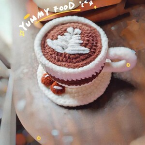 May include: Crocheted coffee cup with a white leaf design on top, sitting on a matching saucer. The cup is brown and white, with a white handle. Two small, brown crocheted cookies are on the saucer. The words "Yummy Food" are above the cup.
