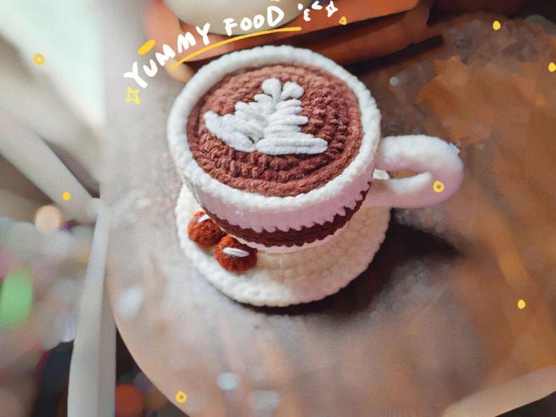 May include: Crocheted coffee cup with a white leaf design on top, sitting on a matching saucer. The cup is brown and white, with a white handle. Two small, brown crocheted cookies are on the saucer. The words "Yummy Food" are above the cup.
