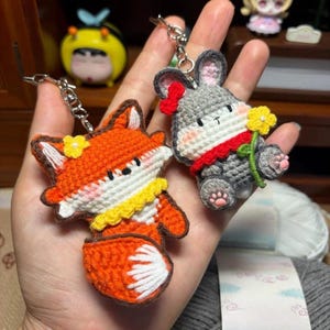 May include: Two crochet keychains featuring a bright orange fox with a yellow collar and a grey rabbit with a red scarf holding a yellow flower. Both figures have embroidered facial features, pink cheeks, and are attached to silver metal key rings.