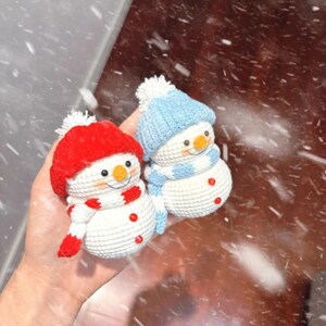May include: Two crocheted snowmen, one with a red hat and scarf, the other with a blue hat and scarf. Both have orange noses, button eyes, and red button accents. The snowmen are handmade and festive.