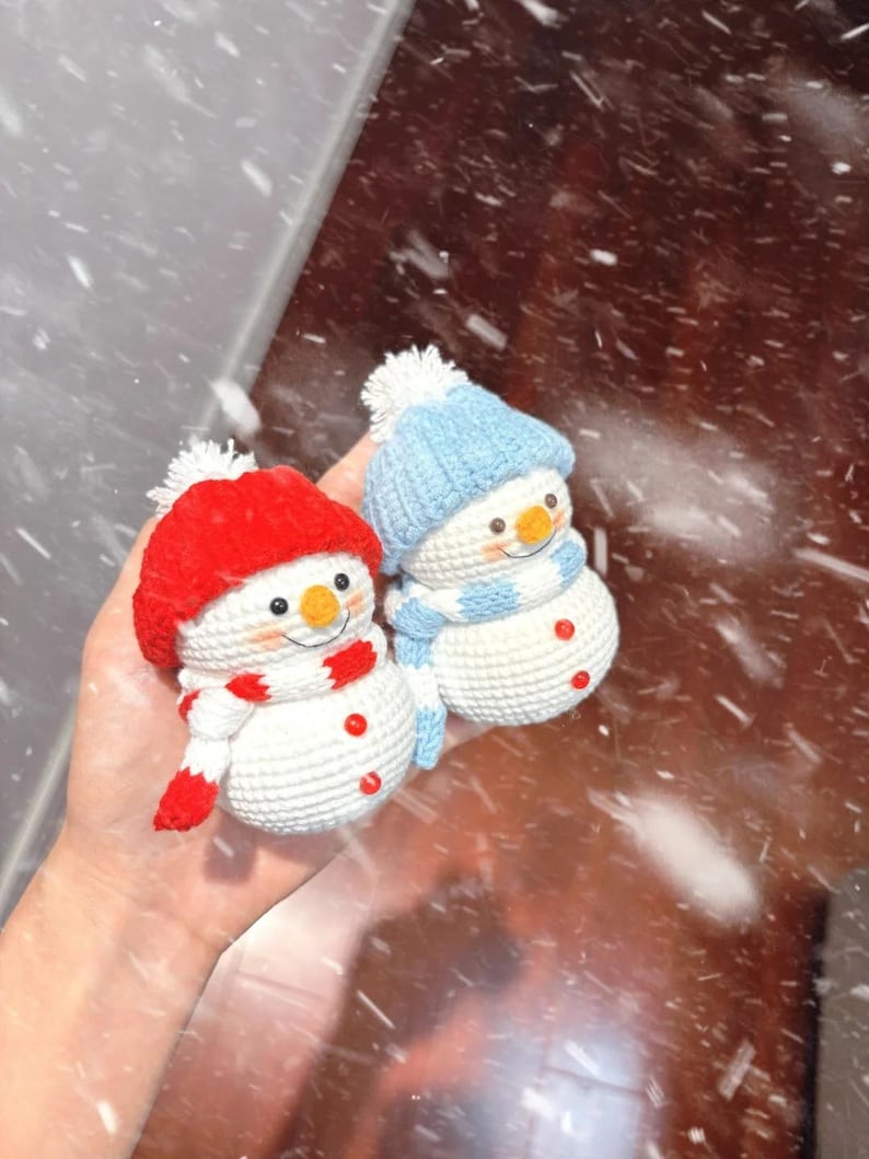 May include: Two crocheted snowmen, one with a red hat and scarf, the other with a blue hat and scarf. Both have orange noses, button eyes, and red button accents. The snowmen are handmade and festive.