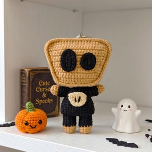 May include: A crochet keychain featuring a square-headed character with a tan face, two black oval eyes, and a black body with a tan chest patch. The character is displayed alongside a small orange crochet pumpkin and a white ceramic ghost figurine.