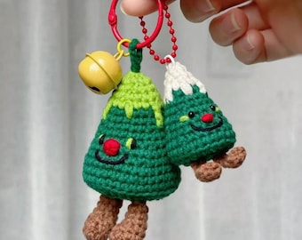 May include: A handmade Christmas tree keychain with a red ring and a yellow bell. The green crocheted trees have smiling faces, red noses, and brown feet. One tree has a white top. A festive holiday accessory.