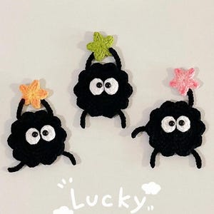 Może przedstawiać: Trzy ręcznie robione szydełkowe stworki z czarnymi ciałami, białymi oczami i małymi ramionami i nogami. Każdy stworek trzyma kolorową gwiazdę nad głową. Poniżej napis "Lucky".