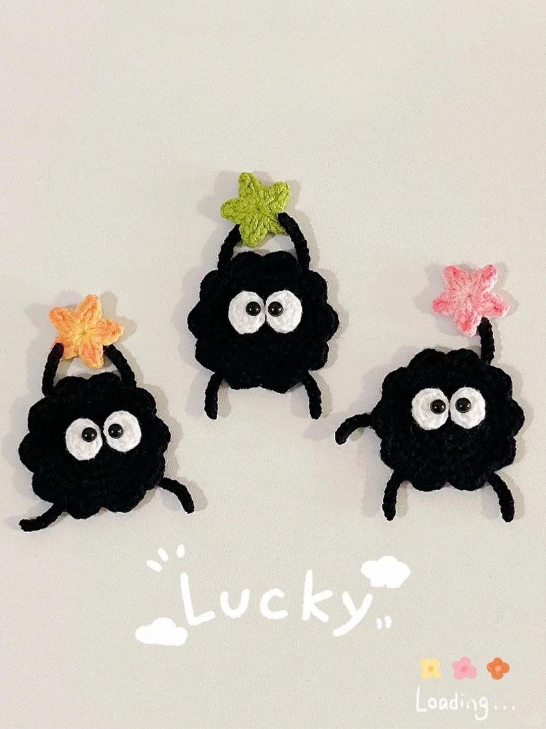 Puede incluir: Tres criaturas hechas a mano con ganchillo, cuerpos negros, ojos blancos y peque&ntilde;os brazos y piernas. Cada criatura sostiene una estrella colorida sobre su cabeza. La palabra "Lucky" est&aacute; escrita debajo.