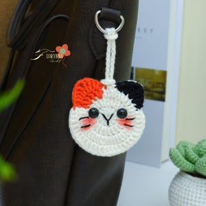 May include: A handmade crochet cat keychain with a white base, orange and black patches, and black button eyes. The cat's face has pink blush and black whiskers. The keychain is attached to a silver ring and a white cord.