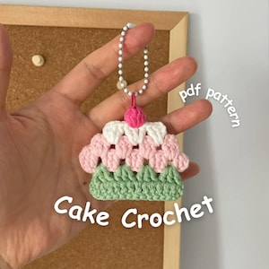 Op de afbeelding: Een gehaakte cake sleutelhanger in roze, wit en groen, met een roze kers erop. De sleutelhanger is bevestigd aan een zilveren kralenketting. De afbeelding bevat de tekst "Cake Crochet" en "pdf pattern".