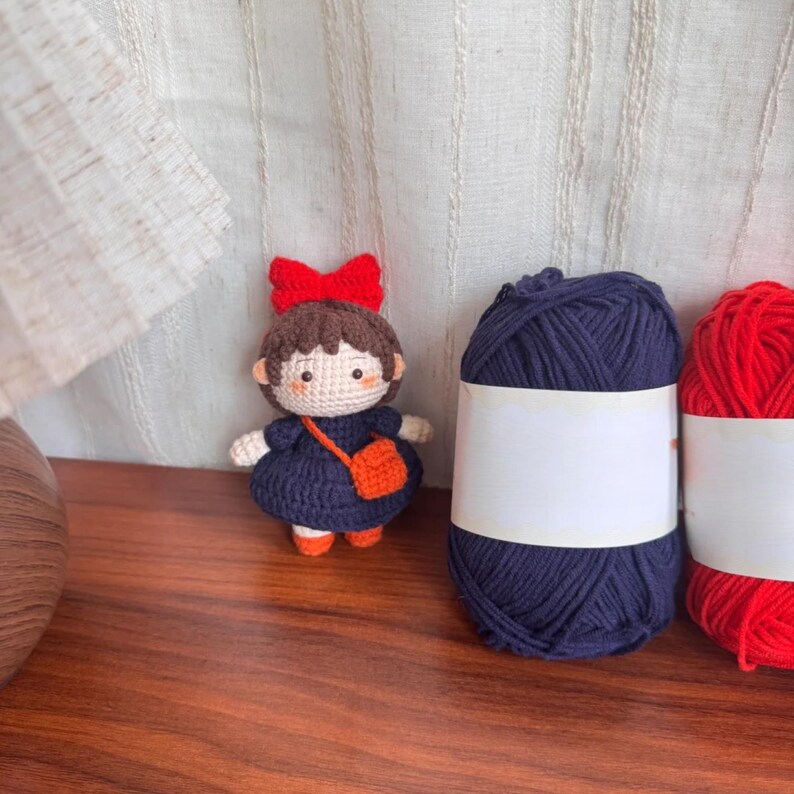 May include: A handmade crochet doll with a red bow, dark blue dress, and orange purse. Next to the doll are two skeins of yarn, one dark blue and one red. The doll is approximately 4 inches tall.