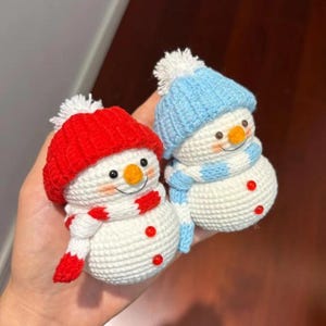 May include: Two handmade crochet snowmen. One snowman wears a red hat and scarf, the other a blue hat and scarf. Both have white bodies, orange noses, and red button accents. The snowmen are a festive winter decoration.