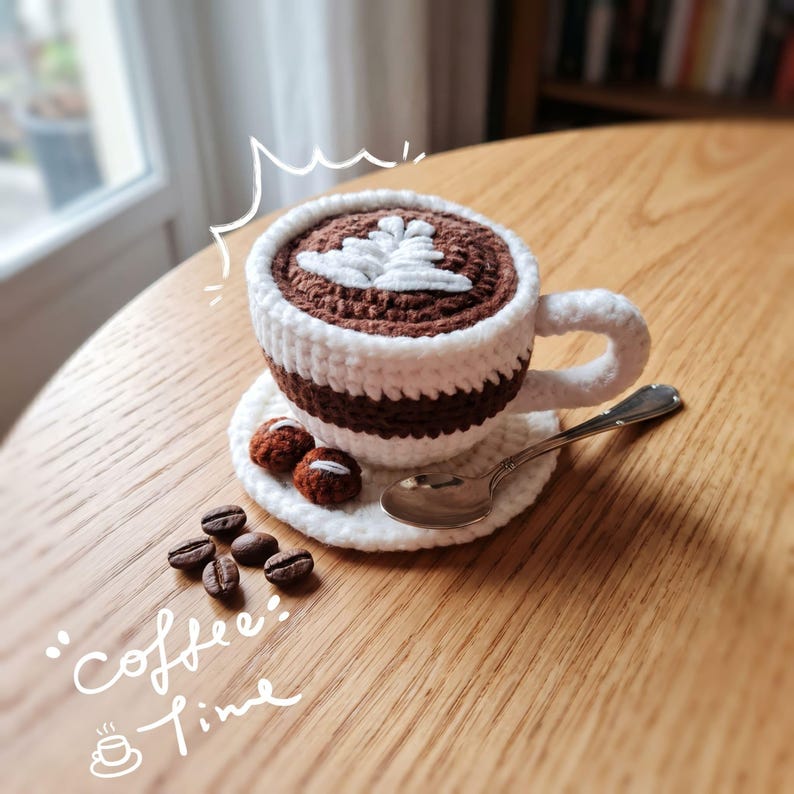 May include: A crocheted coffee cup in white and brown, filled with brown yarn and a white leaf design. It rests on a white saucer with two small crocheted biscuits, a silver spoon, and coffee beans. The words "coffee time" are written on the wooden table.