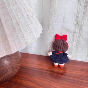 May include: A handmade crochet doll with a dark blue dress, brown hair, and a red bow. The doll has white arms and legs with orange shoes. The doll is standing on a wooden surface.