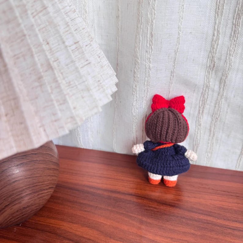 May include: A handmade crochet doll with a dark blue dress, brown hair, and a red bow. The doll has white arms and legs with orange shoes. The doll is standing on a wooden surface.