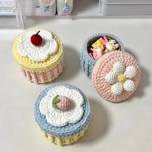 May include: Crocheted cupcake-shaped containers in pastel colors. One has a red cherry, another a strawberry, and a third a flower on top. The containers are open, revealing small toys inside. The containers are handmade.