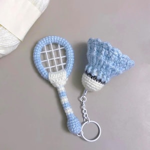 Puede incluir: Juego de llaveros de bádminton de ganchillo con una raqueta y un volante en azul claro y blanco. La raqueta tiene una red blanca y un mango a rayas azules y blancas. El volante es azul claro con base blanca y detalles negros.