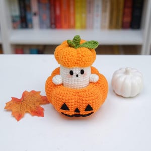 May include: A crocheted white ghost figure wearing an orange pumpkin hat, sitting inside a larger orange crocheted pumpkin with a black felt jack-o'-lantern face. Beside it lies a single orange maple leaf and a small white ceramic pumpkin.
