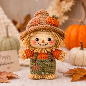 May include: A crocheted scarecrow doll wearing a brown hat with a small orange pumpkin, an orange shirt, and green overalls with brown buttons. The doll has straw-like hair, pink cheeks, and a stitched smile, standing upright as a decorative figure.