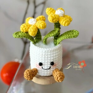 May include: A crocheted white flower pot with a smiling face, holding two yellow flowers with white centers and green stems and leaves. The pot has small brown feet and is sitting on a wooden base.