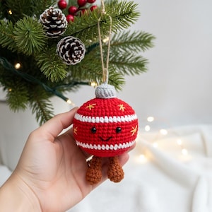 Peut inclure: Une d&eacute;coration de No&euml;l faite &agrave; la main, rouge et blanche, en crochet, avec un visage souriant et de petites jambes marron. L'ornement a des &eacute;toiles jaunes et un dessus gris. Il est suspendu &agrave; une ficelle de ficelle naturelle. L'arri&egrave;re-plan comprend un sapin de No&euml;l.