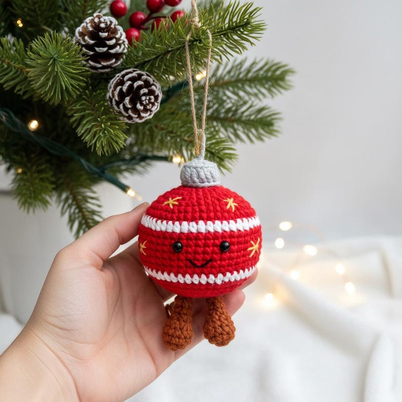 Peut inclure: Une d&eacute;coration de No&euml;l faite &agrave; la main, rouge et blanche, en crochet, avec un visage souriant et de petites jambes marron. L'ornement a des &eacute;toiles jaunes et un dessus gris. Il est suspendu &agrave; une ficelle de ficelle naturelle. L'arri&egrave;re-plan comprend un sapin de No&euml;l.
