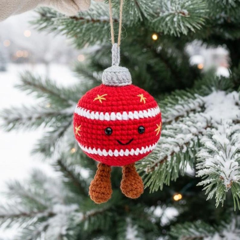 Peut inclure: Une d&eacute;coration de No&euml;l faite &agrave; la main en crochet rouge et blanc, avec un visage souriant, des pieds bruns et des &eacute;toiles jaunes. L'ornement est suspendu &agrave; une ficelle naturelle devant un sapin enneig&eacute;.