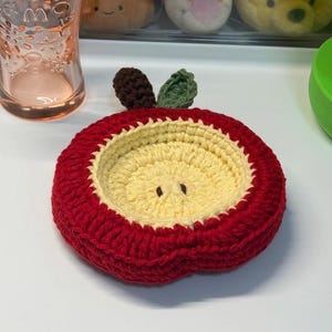 Può includere: Un sottobicchiere a forma di mela all'uncinetto. Il sottobicchiere ha un bordo esterno rosso, un centro giallo e dettagli di foglie marroni e verdi. Il sottobicchiere rotondo è progettato per assomigliare a una mela.