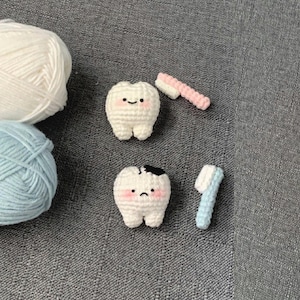 Op de afbeelding: Gehaakte witte tanden met roze wangen en zwarte stippen voor ogen en mond. Eén tand heeft een zwarte vlek. Twee kleine gehaakte tandenborstels, één roze en één blauw, zijn ook aanwezig. Twee bollen wol staan op de achtergrond.
