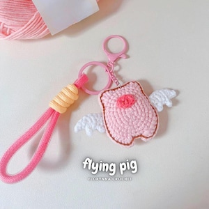 May include: A handmade pink crochet keychain shaped like a pig with white wings. The keychain has a pink strap and a pink clasp. The text "flying pig" and "FLORYNNA CROCHET" are printed on the bottom of the image.