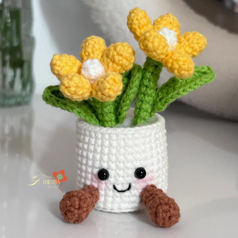 May include: A handmade crochet flower pot with yellow flowers, green leaves, and a white pot. The pot has a smiling face with black eyes and a curved mouth. The pot has brown feet.