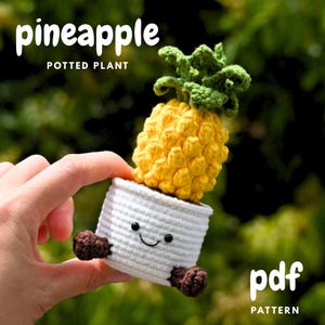 May include: A crocheted pineapple potted plant with a smiling face on the white pot. The pineapple is yellow with green leaves. The text "pineapple potted plant" is at the top of the image, and "pdf pattern" is at the bottom.