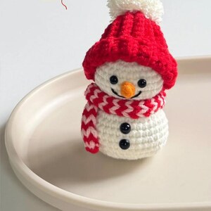 May include: A handmade crochet snowman figurine. The snowman has a white body, a red hat with a white pom-pom, and a red and white scarf. It features black button eyes, a carrot nose, and a smiling mouth. A festive winter decoration.
