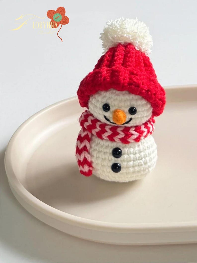 May include: A handmade crochet snowman figurine. The snowman has a white body, a red hat with a white pom-pom, and a red and white scarf. It features black button eyes, a carrot nose, and a smiling mouth. A festive winter decoration.
