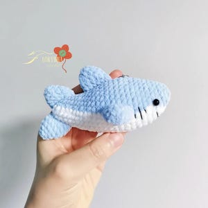 May include: A handmade, light blue and white crocheted shark toy. The shark has black bead eyes and black stitched gills. The toy is held in a hand against a white background. The shark is a cute, soft toy.