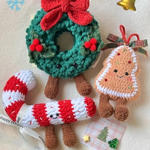 以下が含まれることがあります： 手作りのクリスマスかぎ針編みオーナメントのコレクション。赤いリボンとベリーが付いた緑色のリース、キャンディケーン、ジンジャーブレッドツリーが含まれています。各オーナメントには笑顔の顔と茶色の脚が付いています。「Merry Christmas」と書かれたカードも表示されています。