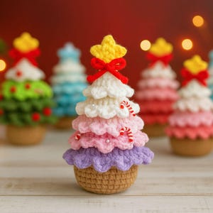 Puede incluir: Árbol de Navidad tejido a ganchillo con una estrella amarilla y un lazo rojo. El árbol presenta capas de blanco, rosa y morado, adornado con bastones de caramelo. Se asienta sobre una base tejida a ganchillo marrón, ideal para la decoración navideña.
