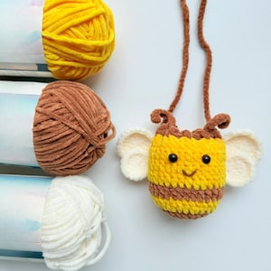 May include: A handmade crochet bee-shaped purse with a brown strap, antennae, and stripes. The bee has black eyes and a smiling mouth. The purse is yellow and brown, with white wings. Beside the purse are three balls of yarn in yellow, brown, and white.