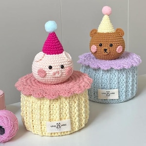 May include: Two handmade crochet storage containers. One is yellow with a pink pig wearing a pink party hat and a blue pom-pom. The other is blue with a brown bear wearing a yellow party hat and a pink pom-pom.