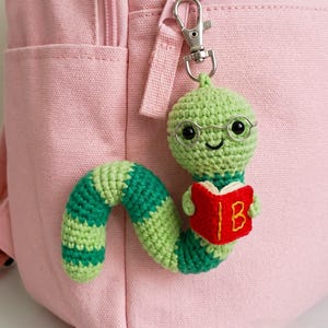 May include: A crochet keyring featuring a green worm with a striped body, wearing thin wire-rimmed glasses and holding a small red book with the letter B embroidered in yellow on the cover. The worm is attached to a silver metal lobster clasp.