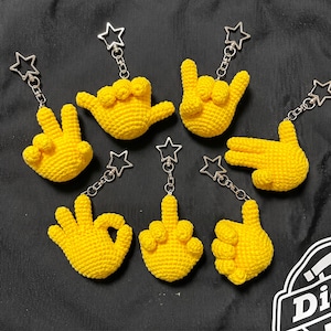 Puede incluir: Una colección de siete llaveros de ganchillo amarillo con gestos de mano, cada uno con un cierre plateado en forma de estrella. Las formas de las manos incluyen un signo de paz, un signo shaka, un signo de rock, un dedo señalando, un signo de OK, un dedo medio y un puño.