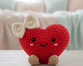 May include: A handmade, red crocheted heart-shaped plush toy with a cream-coloured bow. The toy has black eyes, rosy cheeks, and small tan feet. The plush is sitting on a light-coloured wooden surface.