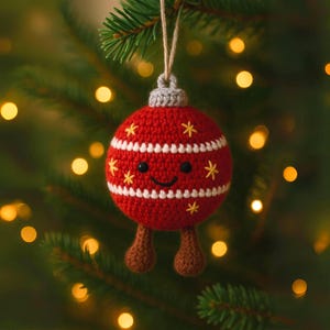 Peut inclure: Une boule de No&euml;l rouge crochet&eacute;e avec des rayures blanches et des &eacute;toiles jaunes. L'ornement a un visage souriant et des jambes crochet&eacute;es marron, suspendu &agrave; un sapin de No&euml;l vert. La boule mesure environ 7.6 cm de diam&egrave;tre.