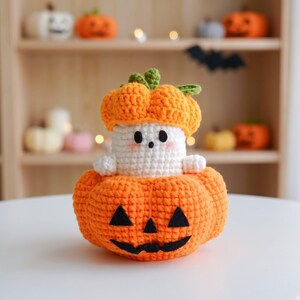 May include: A crocheted ghost figure wearing an orange pumpkin hat, sitting inside a larger orange crocheted pumpkin with a black felt jack-o'-lantern face. The ghost has a white body, two black eyes, a small black mouth, and pink blush cheeks.