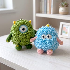 May include: Two round, textured crochet plush keychains. The larger one is green with a single central eye and small yellow horns. The smaller one is light blue with two eyes and pink arms and feet. Both feature metal key rings on top.